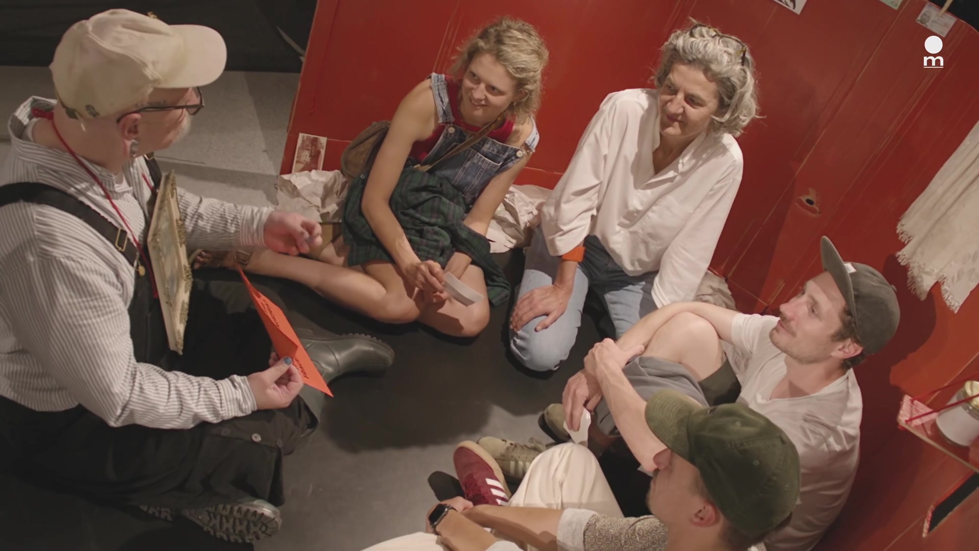 Participants seated in a circle and exchanging stories against the red set of May I Stay?, in a still from the Mozarteum trailer.